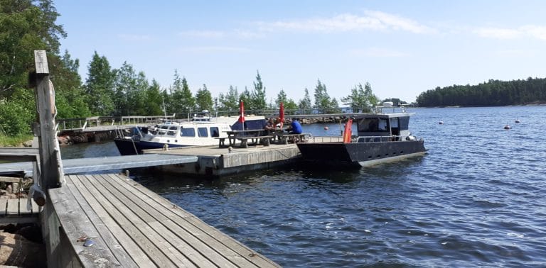 Laiturissa kaksi venettä ja ihmisiä istumassa pöydän ääressä. Taustalta pilkistää kivistä tehty aallonmurtaja, jonka päällä kasvaa puita.