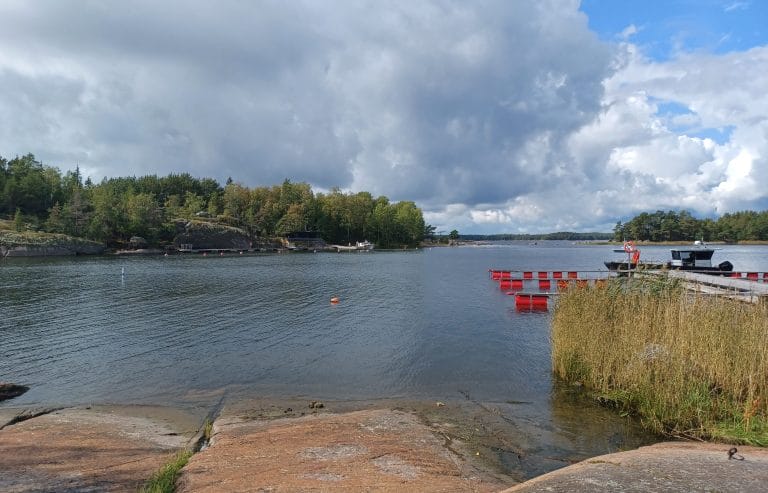 Näkymä Sisä-Nuokolta Ulko-Nuokon laiturille päin. Etualalla kallio ja oikealla puolella laiturin punaiset ponttoonit ja ruovikkoa. Taivaalla tummia pilviä.