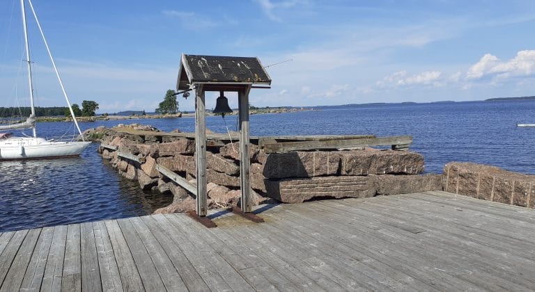 Laiturilla pienen katoksen alla oleva valettu kello ja kivistä tehty aallonmurtaja, jossa kiinnittyneenä purjevene. Näkymä merelle päin.