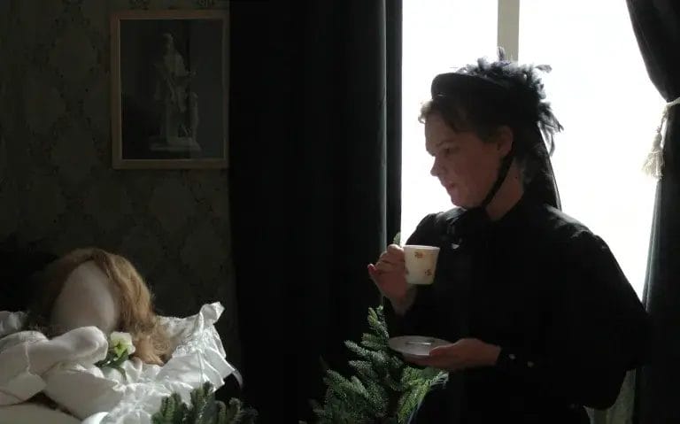 Nainen mustassa puvussa juo kahvia ja suree nukkevainajaa.