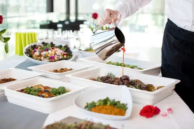 Tarjoilulautasia täynnä ruokaa ja kädellä kaadetaan lastikekannusta kastiketta kulhoon.