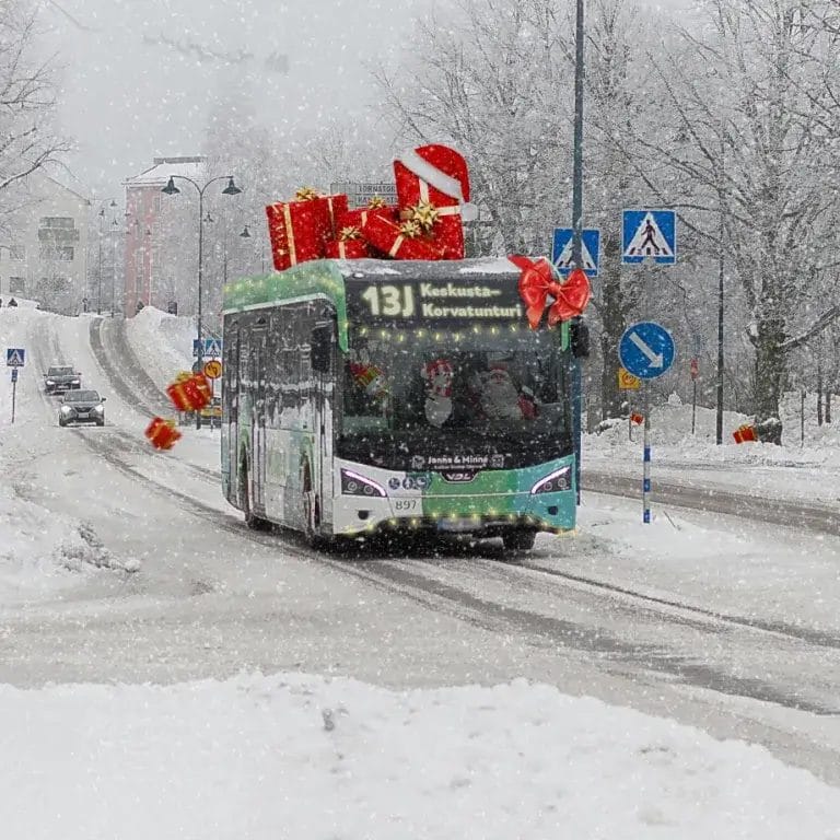 Linja-auto ajamassa lumisella kadulla, mikä on koristeltu paketein ja sitä ajaa joulupukki.