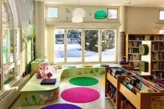 Kirjaston lastenosasto, jossa isot ikkunat, hyllyjä ja värikkäitä mattoja.